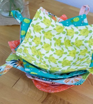 A stack of fabric bowl cozies in coral, teal and citrus prints with fabric loop handles, on a wooden table