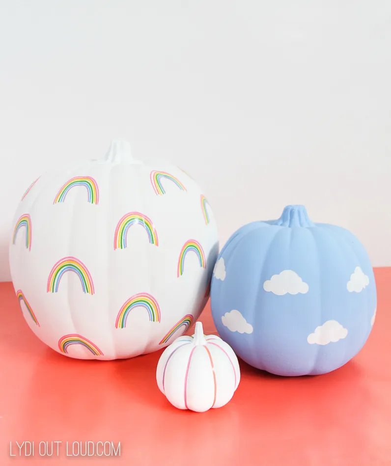 Three faux pumpkins decorated with vinyl decals: a large white one with rainbows, a blue one with white clouds and a small white one with thin coloured stripes