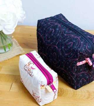 Two quilted boxy zip pouches, a large navy and pink patterned one and a smaller cream bicycle print one, on a wooden table