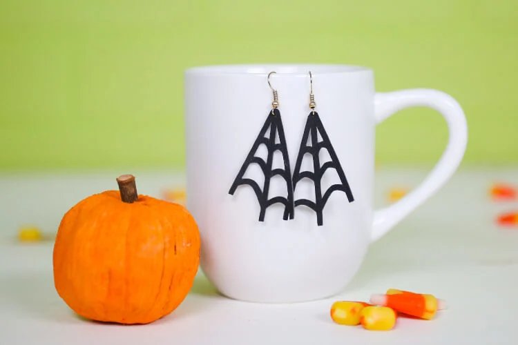 Black faux suede spider web earrings hanging from a white mug beside a small pumpkin and candy corn