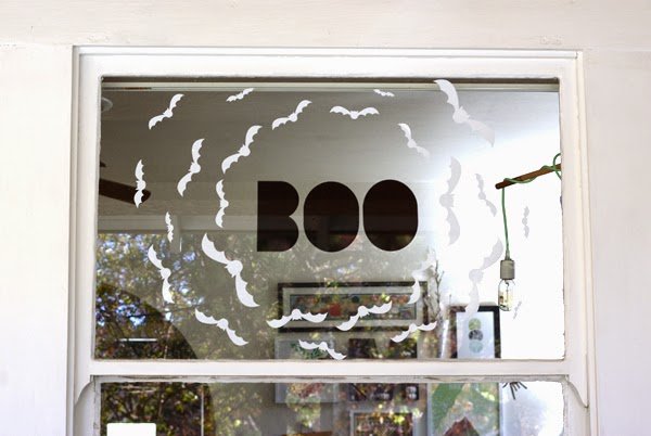 White paper bats and a black BOO sign stuck to a glass door window, with a room and hanging light bulb visible through it