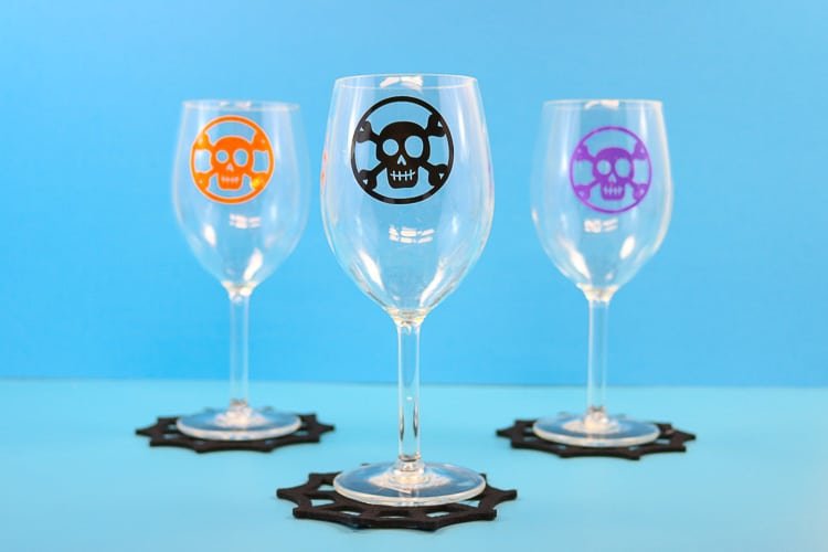 Three wine glasses on a blue background with orange, black and purple skull and crossbones decals, standing on black spider web coasters