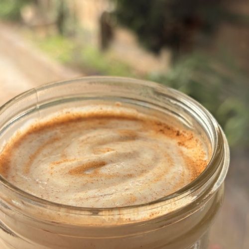 A hand holding a glass jar of creamy chicha venezolana con pasta with a cinnamon swirl on top, in a sunlit garden