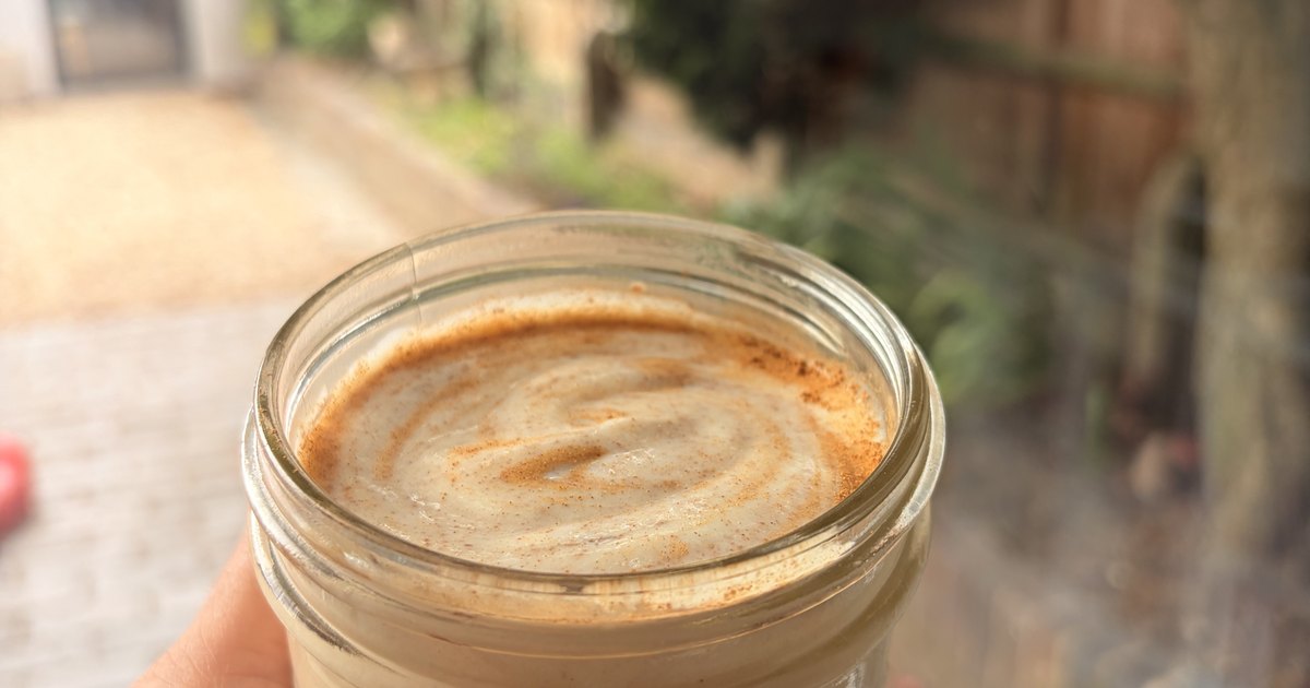 A hand holding a glass jar of creamy chicha venezolana con pasta with a cinnamon swirl on top, in a sunlit garden