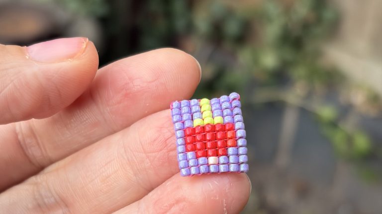 A handmade square-stitch beaded pendant made from cylinder beads, held between finger and thumb outdoors